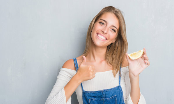 Beautiful young woman over grunge grey wall holding a lemon slice happy with big smile doing ok sign, thumb up with fingers, excellent sign