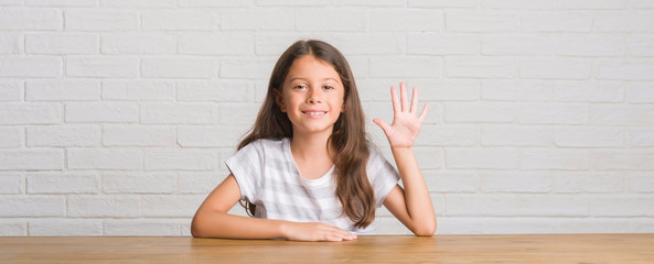 Young hispanic kid sitting on the table at home showing and pointing up with fingers number five...