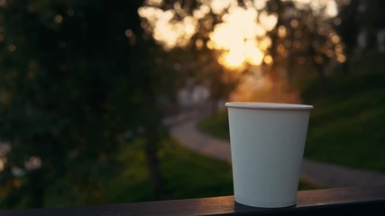 A white paper cup with coffee or tea stands on the railing against the backdrop of the city park at sunset. A hot drink raises steam. - Powered by Adobe