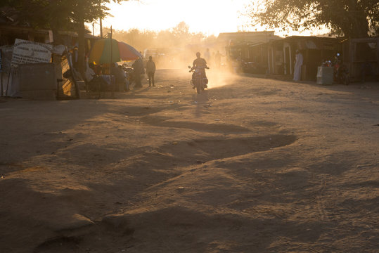 A motorbike rides forward leaving behind a cloud of dust in the air which gets highlighted by the grazing sunlight  of a late afternoon
