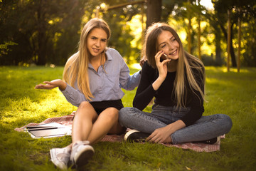 Displeased confused girl looking at camera near her friend sister talking by mobile phone students sitting in the park outdoors on grass.
