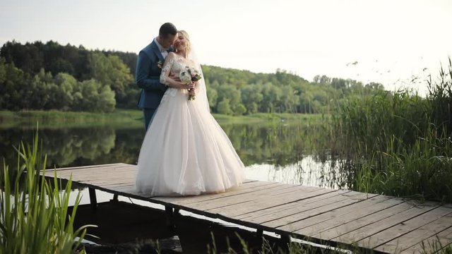 Smiling beautiful young wedding couple stand hugs near lake love bride dress groom outdoors white married woman celebration romantic beauty female man people suit husband kiss slow motion