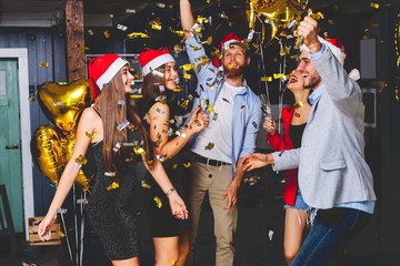 Celebrating New Year together. Group of beautiful young people in Santa hats throwing colorful confetti and looking happy.