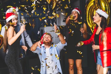 Celebrating New Year together. Group of beautiful young people in Santa hats throwing colorful confetti and looking happy.