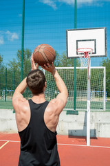 Back view portrait of muscular young man throwing ball in hoop while playing basketball in outdoor...