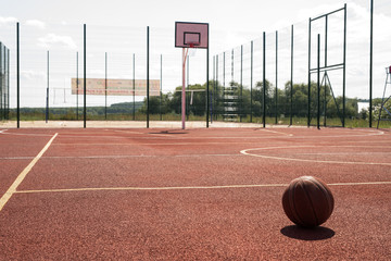 Fototapeta premium Wide angle shot of empty outdoor basketball court lit by sunlight with hoop and ball on floor in foreground, copy space
