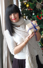 Christmas. A happy smiling woman in a warm scarf is holding a present. Against the backdrop is a dressed fir.