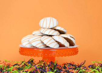 Orange pedestal with pumpkin muffin top cake cookies frosted, orange, black and green shredded paper below, orange background with copy space. Thanksgiving or Halloween theme.