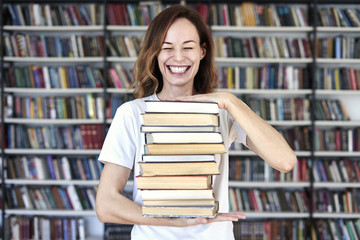Woman model college student with books at library holds bunch of books, looks smart, smiling to camera. bookshelves at the library. Knowledge and self-development