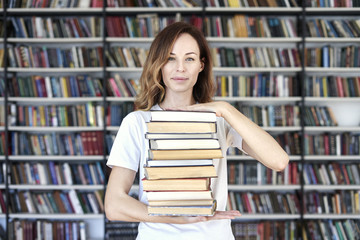 Woman model college student with books at library holds bunch of books, looks smart, smiling to camera. bookshelves at the library. Knowledge and self-development