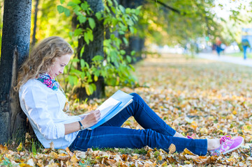 Teen student woman sitting on lawn in park with books and notebook and writes