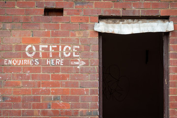 Old painted sign, Office Enquiries Here, on a red brick wall and an open doorway