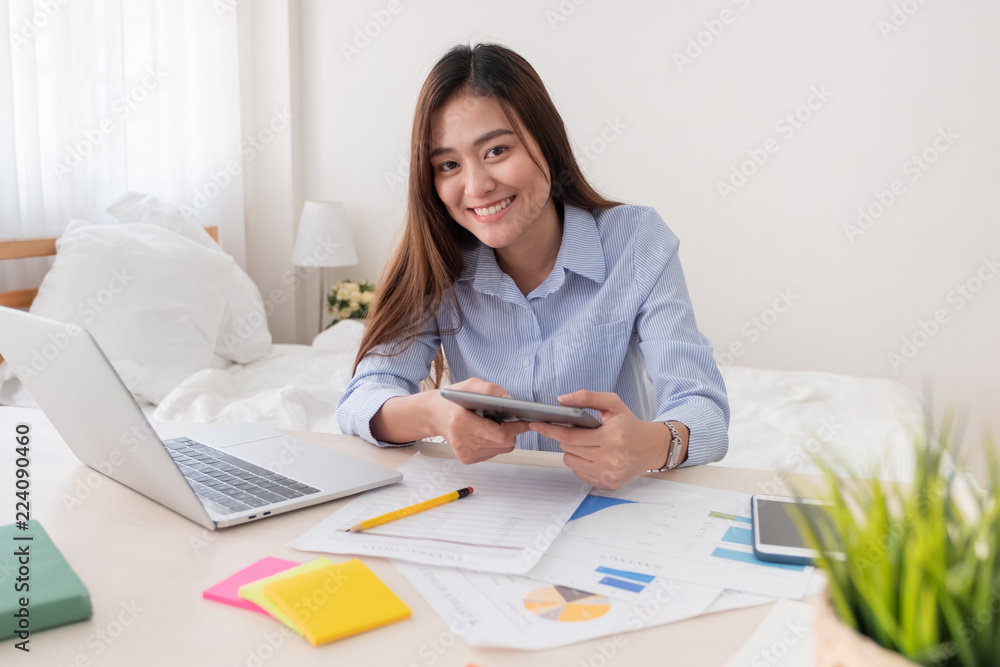Poster asian freelancer looking at camera and holding tablet while working with laptop on table in bedroom 