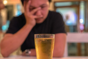 Asian young man in lonely and depressed action and holding face in hands and see glass of beer in pub and restaurant with low light place, depression and drinking concept