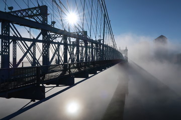 An almost silhouette of a beautiful bridge stands over a thick morning fog covering the river water below as the suns reflection tries to shine through but succeeding up above in the blue sky.