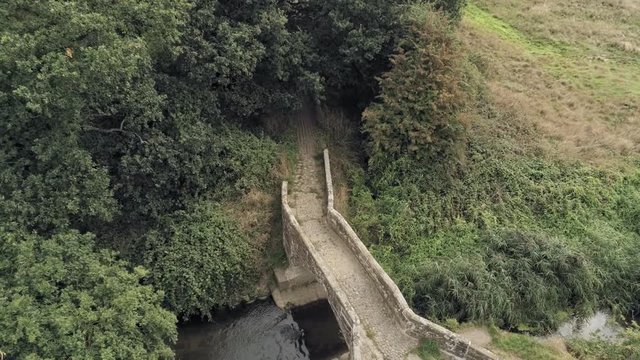 Aerial view, down move. Old small stone roman bridge and grass, overgrown water stream in english countryside.
