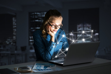 Attractive blonde wearing glasses in dark office using laptop. Mixed media