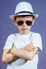 A little boy in a white hat and t-shirt in sunglasses on a purple background. The child smiles and rejoices.
