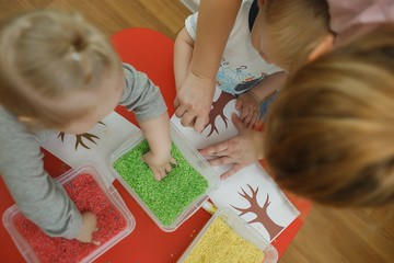 Group of children playing with colored rice from the sensory box. Painting exercise. Baby's sensory educational kit - obrazy, fototapety, plakaty
