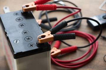 A Car battery with red and black battery Jumper Cables with copper clamps attached to the terminals. Automotive battery on a work desk with tools in background.