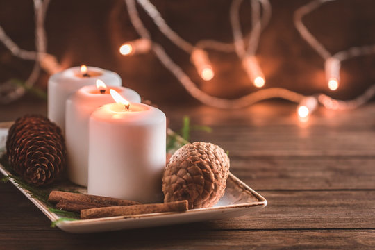 Winter cozy decoration with white burning candles on a wooden  festive table and blurrred lights on a background. Warm toned image.