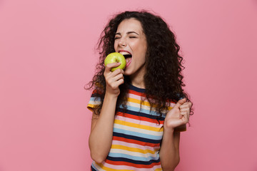 Photo of beautiful woman 20s with curly hair eating green apple, isolated over pink background - obrazy, fototapety, plakaty