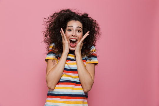 Image of surprised woman 20s with curly hair smiling and touching face, isolated over pink background