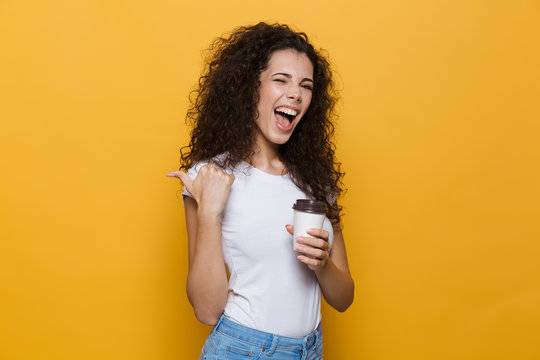 Image of pretty young woman 20s with curly hair smiling and holding takeaway coffee in paper cup, isolated over yellow background