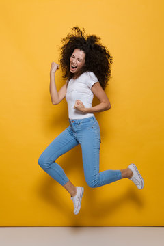 Full length photo of young woman 20s with curly hair having fun and jumping, isolated over yellow background