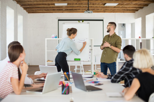 Young man in eyeglasses and shirt and pretty woman in blouse standing near board happily presenting new project to colleagues. Group of creative people working together in modern office