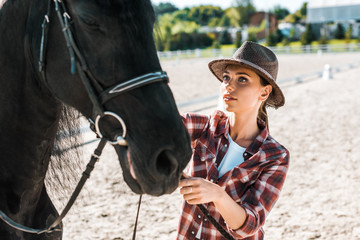 Obraz premium attractive jockey in checkered shirt and hat fixing horse halter at ranch