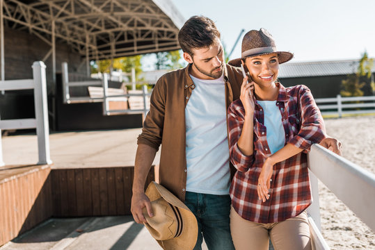 stylish cowboy and cowgirl in casual clothes standing near fence at ranch, woman talking by smartphone