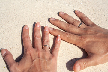 Tanned hands of newlyweds with gold rings on ring fingers on white sand as a symbol of wedding...