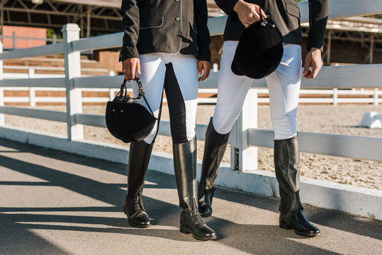 cropped image of equestrians in professional apparel walking near fence and holding riding helmets at ranch
