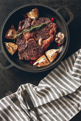 elevated view of cooked steak with lemons and berries on frying pan, striped tablecloth on surface in kitchen