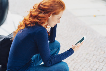 rothaarige frau sitzt draußen auf einer treppe und schaut auf ihr mobiltelefon
