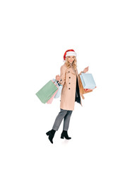 full length view of beautiful happy young woman in santa hat holding shopping bags and smiling at camera isolated on white