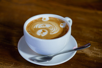 a white cup with latte and teaspoon stand on the platter on the table in the cafe. Hot latte with a stencil in the form of a bear cools in the cup