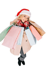high angle view of happy young woman in santa hat and mittens holding shopping bags and smiling at camera isolated on white