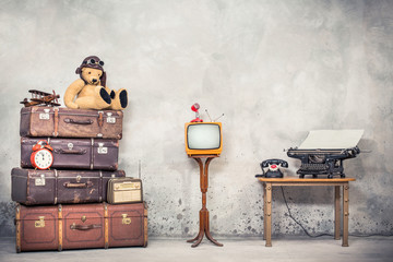 Teddy Bear toy with leather aviator's hat and goggles sitting on retro old  travel suitcases, wooden plane, alarm clock, TV, mic, telephone and typewriter. Loft storage. Vintage style filtered photo