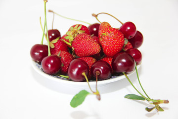 red berries of strawberries and burgundy cherries with green leaves on a plate isolated on white background