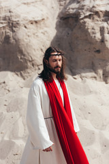 handsome Jesus in robe, red sash and crown of thorns walking with closed eyes in desert