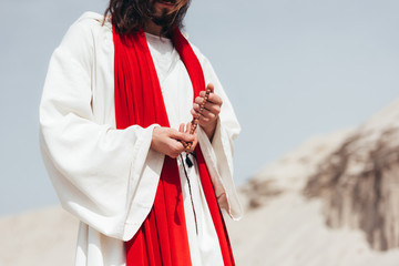 cropped image Jesus holding wooden rosary and praying in desert