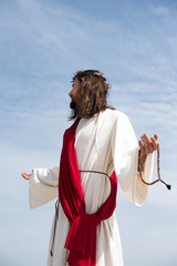 Jesus in robe, red sash and crown of thorns holding rosary and standing with open arms against blue...