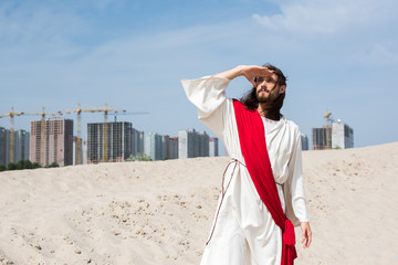 Jesus in robe, red sash and crown of thorns standing on sand and looking up with buildings on...