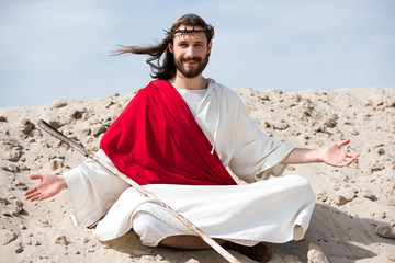 smiling Jesus in robe, red sash and crown of thorns sitting in lotus position with open arms on...