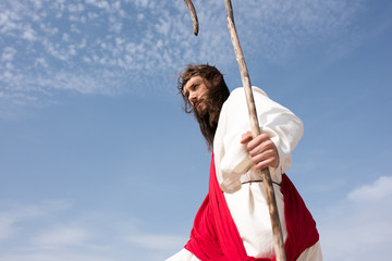low angle view of Jesus in robe, red sash and crown of thorns standing with staff against cloudy sky