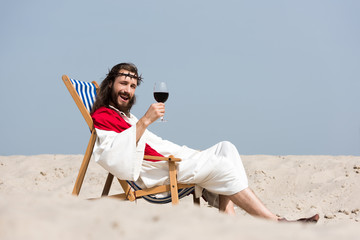 smiling Jesus in robe and red sash resting on sun lounger with glass of red wine in desert, looking at camera