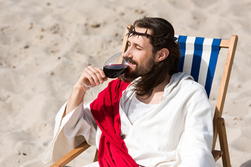Jesus in robe and red sash resting on sun lounger and drinking red wine in desert
