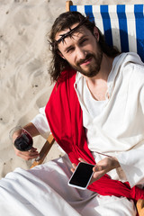 Jesus resting on sun lounger with glass of wine and holding smartphone with blank screen in desert